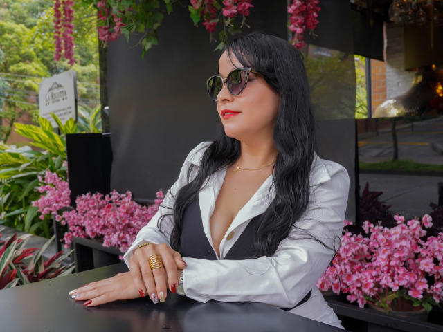 TamaraFoxy, con i capelli neri, il rossetto rosso e gli occhiali da sole, indossa una camicia bianca con un gilet nero, appoggiata a un tavolo, circondata da fiori rosa. Il seno è visibile, indossa un anello d'oro..