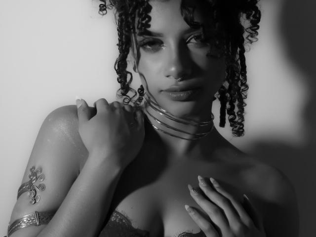 NoemiDawson with curly hair, wearing a necklace, touching her chest, hand on collarbone, showing a tattoo on her upper arm, and a nose ring, in black-and-white.