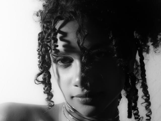 NoemiDawson's face, dark curly hair, shadowed eyes, subtle smile, thick choker, black and white, high-contrast, intense gaze, soft shadows on her skin, sensual, intimate, detailed.