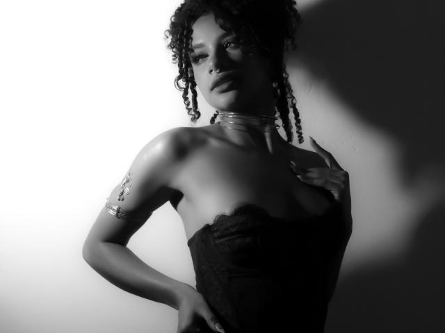 NoemiDawson with dark curly hair, wearing a strapless black lace dress, shiny choker, and arm bracelet, hand on chest, black and white, shadow on wall, looking seductive.
