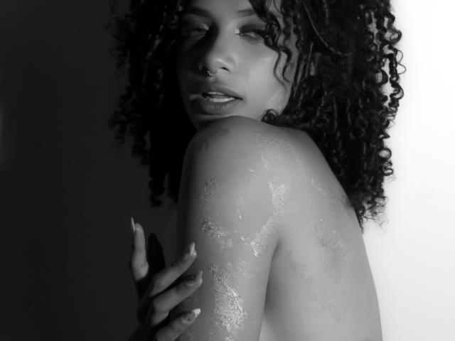 Black-and-white photo of NoemiDawson with curly hair, nose ring, and shiny, oily shoulder, looking seductively at the camera, holding her arm, showing off her tits.