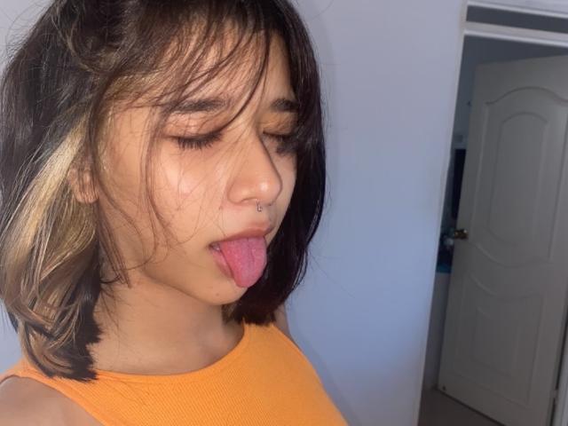 LoveLali with dark brown hair and blonde highlights, nose ring, and orange top, tongue out, eyes closed, in a selfie. Her ass is not visible.
