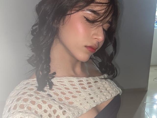 LoveLali with wavy black hair, nose ring, and pinkish blush, wearing a crocheted off-shoulder top, eyes closed, looking down, soft light on her face, showing her tits.