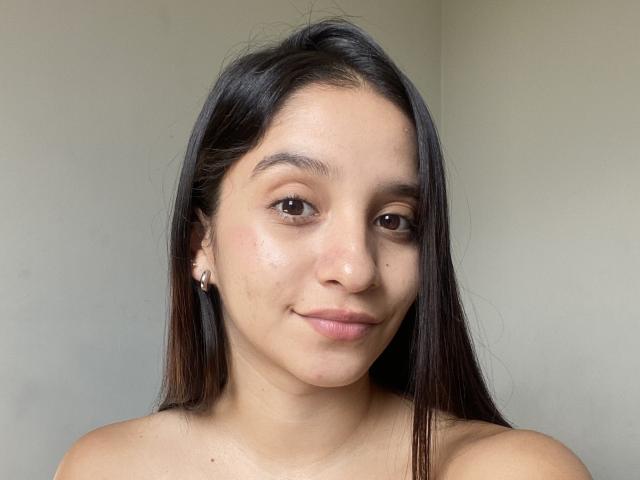 MabelPrado with long dark hair, brown eyes, and a subtle smile, showing her bare shoulders, wearing small earrings, and a slight nose freckle, in a neutral light.