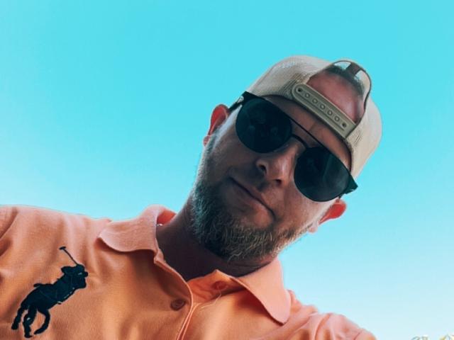 MikeHottLover69, a bearded dude with sunglasses, a beige cap, and an orange polo with a black horse logo, against a bright blue sky.