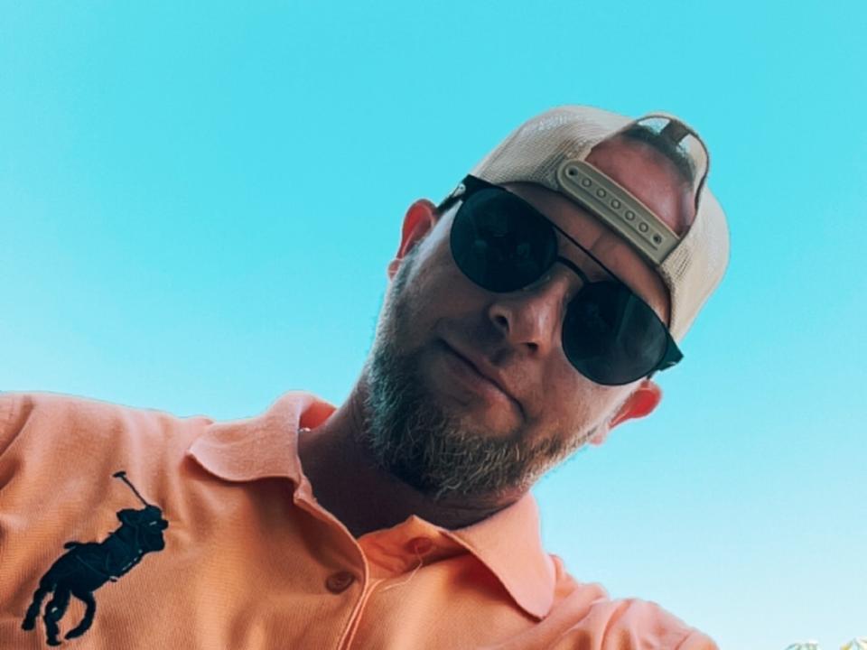 MikeHottLover69, a bearded dude with sunglasses, a beige cap, and an orange polo with a black horse logo, against a bright blue sky.