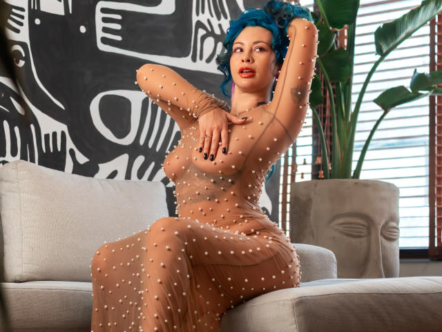 KaatArt with blue hair, wearing a sheer, bead-covered dress, sits on a grey couch, arms raised, showing her tits and red lipstick, in a brightly lit room.