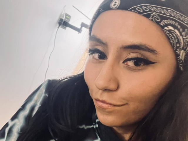 EviieGothel has brown eyes, a nose ring, and a black bandana with white patterns. She's wearing a black tie-dye shirt, with long dark hair and winged eyeliner.