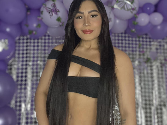 AshlyFerria, con cabello largo y negro, vestida con una blusa negra con un hombro descubierto que muestra sus grandes senos, sonriendo con lápiz labial rojo, rodeada de globos morados y un fondo a cuadros..