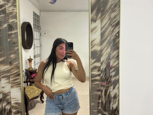 SharonKlart, con cabello negro largo, rostro rosado, una blusa blanca sin mangas y pantalones cortos vaqueros de cintura alta, tomando una selfie frente a un espejo, sosteniendo un teléfono negro, en una habitación..