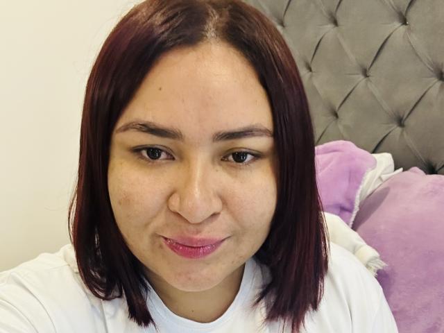 KarlaWild with medium brown skin, red-brown straight hair, small nose, slight smile, pink lipstick, white shirt, and freckles, sitting against a grey tufted headboard.