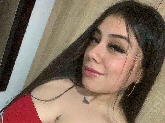 KatalinaSexy69 has long black hair, thick eyelashes, pink glossy lips, hoop earrings, and a small butterfly tattoo below her neck. She's wearing a red top, showing her tits.