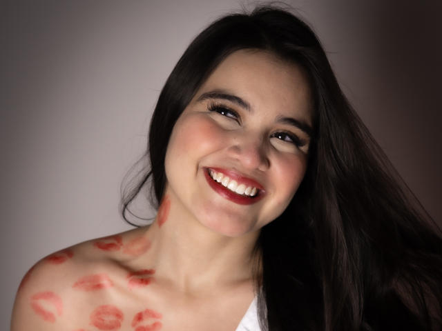 CamilaSofiia has dark hair, red lipstick, and a smile showing white teeth. She has red lipstick kisses on her bare shoulder and upper chest. Her eyes are brown.
