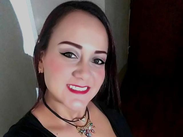 DivaCurvy has dark brown hair, red lipstick, black eyeliner, and is wearing a black top with a necklace. Her tits are visible, slightly highlighted by the lighting.