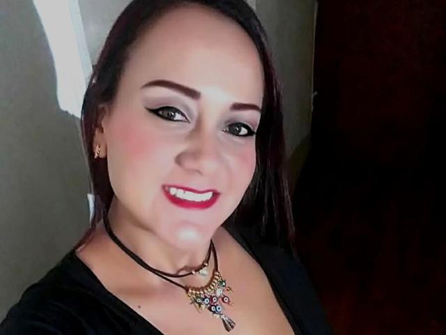 DivaCurvy has red lipstick, black eyeliner, and a smile. She's wearing a black necklace with colorful charms and a black top. Her dark brown hair is straight.