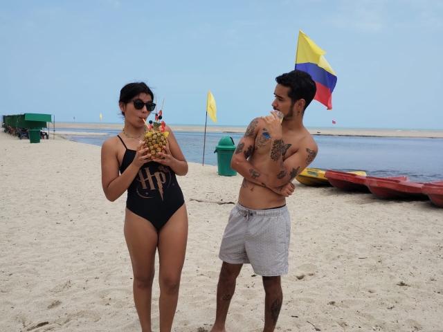 LailaAsher com um maiô preto de peça única, segurando uma bebida de coco, e LailaAsher com tatuagens, shorts cinzas, em pé na praia, olhando uma para a outra. LailaAsher tem seios pequenos, cabelo escuro..