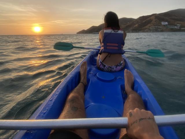 LailaAsher's ass in purple floral shorts, wearing a blue life jacket, paddles in blue kayak, sun setting over ocean, LailaAsher's legs in foreground, sunlit water.