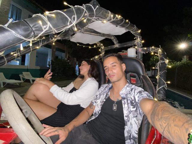 LailaAsher with tits in a black dress, sitting in a car with a dude in a black shirt, dog tags, and a tattooed arm, under a string light canopy.