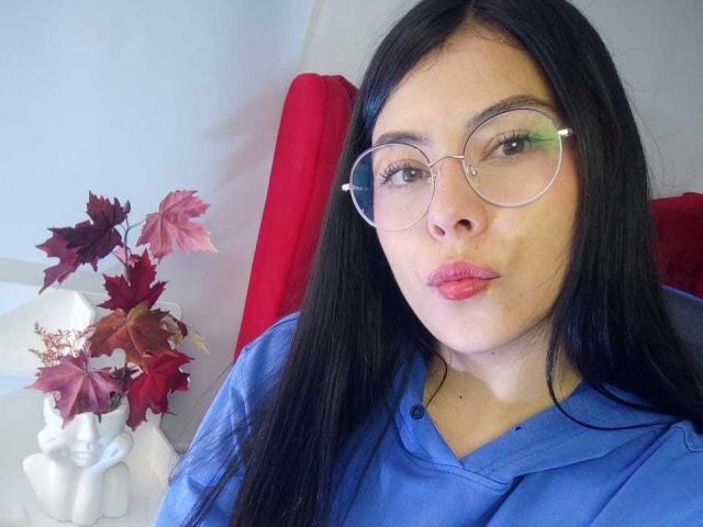 Sophia Ramirez, con cabello negro largo, usando gafas redondas, una sudadera azul y lápiz labial rosa, sentada en una silla roja, con un jarrón blanco lleno de hojas rojas y rosas..