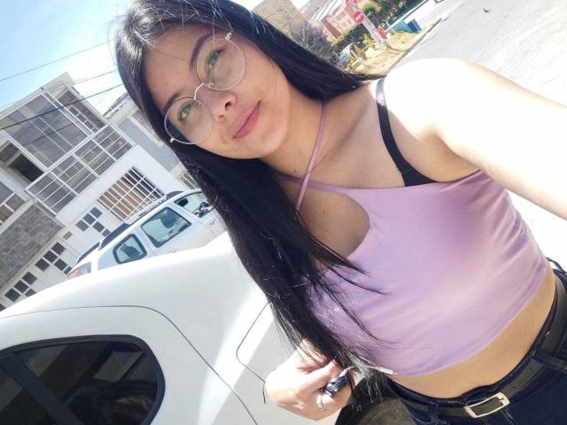 SophiaRamirez with long black hair, glasses, purple crop top, black bra visible, and jeans, standing by a white car, sunlight highlighting her face, and a slight smile.