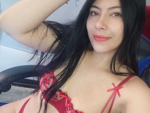 Sophia Ramirez, con cabello negro largo, un sujetador de encaje rojo y un lazo rosa, sentada, con el brazo levantado, mostrando senos pequeños, piel clara, lápiz labial rosa, una ligera sonrisa..