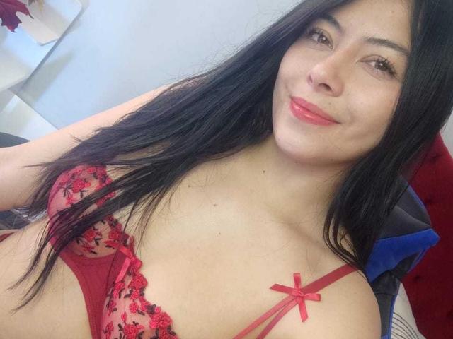 SophiaRamirez with long black hair, red lipstick, and a cute smile, wearing a red lace bra with black floral embroidery and a small red bow on the left cup.