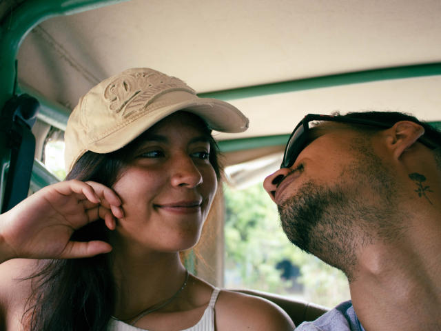 LailaAsher with tan cap, brown hair, smiling at bearded man with tattoo on neck, wearing sunglasses, inside vehicle. Her hand rests near cheek, his head leans in.