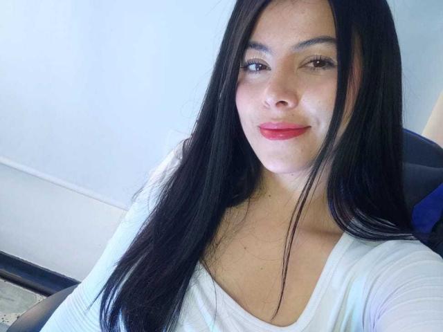 SophiaRamirez tiene el pelo largo y negro, piel clara y lápiz labial rojo. Lleva una blusa blanca, está sentada en una silla y sonríe ligeramente. Sus pechos son pequeños y su vulva es invisible..