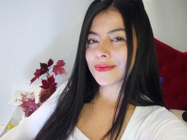 SophiaRamirez tiene el pelo largo y negro, lápiz labial rojo y una blusa blanca. Está sonriendo suavemente, y sus senos son ligeramente visibles. Sus ojos marrones están relajados, y es linda..