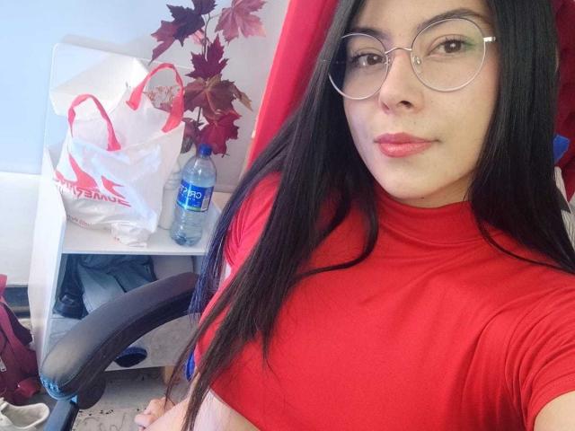 SophiaRamirez with long black hair, wearing round glasses and a red turtleneck, showing her small tits in a selfie, sitting on a red chair with a water bottle.