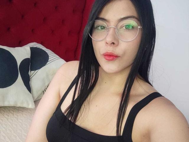SophiaRamirez with black hair, glasses, green-lensed glasses, red lipstick, black tank top, showing her tits, red tufted headboard, white pillow with black circles, light skin.