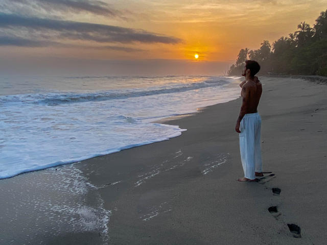 LailaAsher, zonder shirt, met een gespierd lichaam, witte broek, staand op een zandstrand, kijkend naar een zonsondergang, golven die de kust raken, donkere huid, zonder schoenen, bomen op de achtergrond..