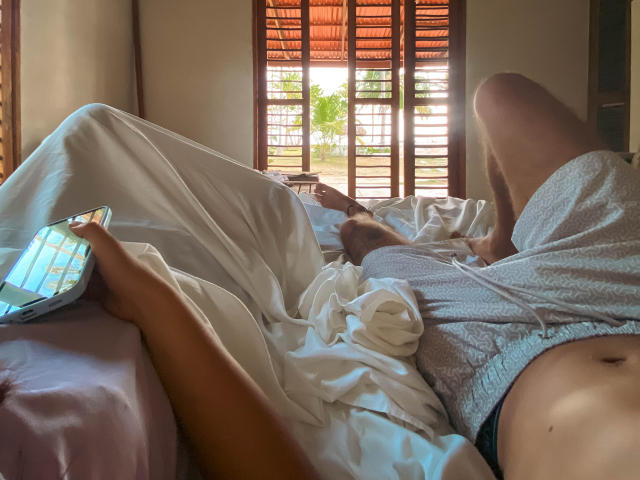 LailaAsher is lying on a bed, wearing grey shorts, with white sheets, holding a phone, and her legs are up, showing her bare ass and small tits.