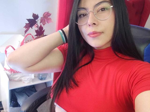 SophiaRamirez with long black hair, red t-shirt, and glasses, showing off her small tits and pink lips, resting her arm on a chair.