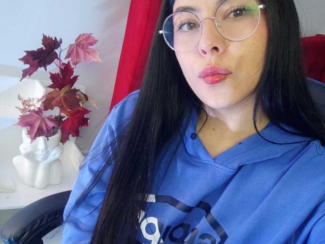 SophiaRamirez with long black hair, glasses, pink lipstick, and blue shirt, pouting her lips, sitting in chair, with red and purple leaves in white vase on left.