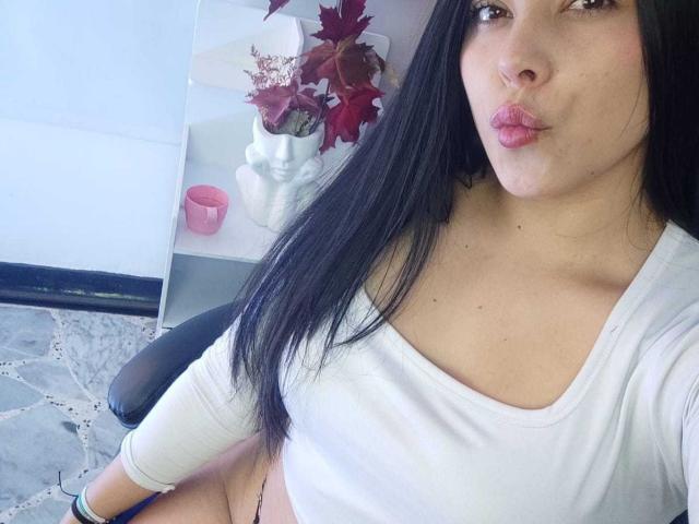 SophiaRamirez with pouty lips, black hair, white long-sleeve top, exposing her small tits, sitting on a chair, wearing a black bracelet, and a white vase in the background.