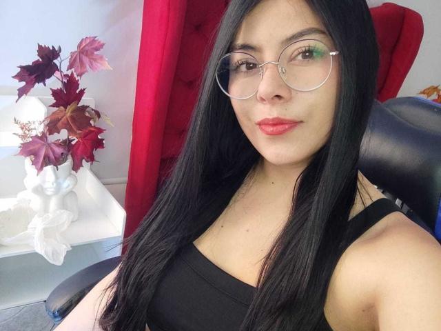 SophiaRamirez with long black hair, wearing round glasses and a black tank top, sitting in a red chair, showing red lips and a subtle smile, with a small vase.