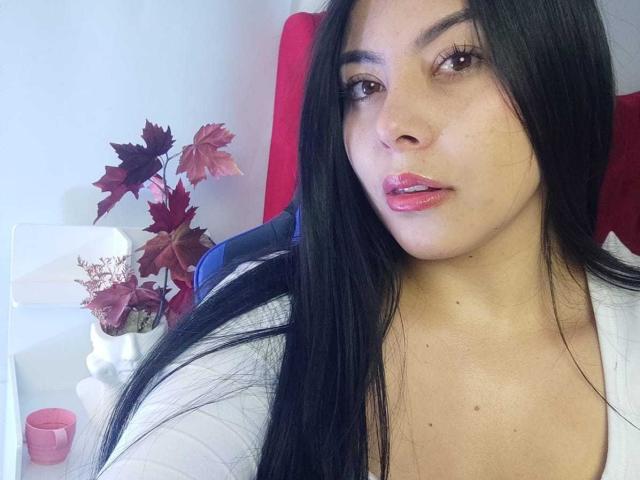 SophiaRamirez with long black hair, brown eyes, and pink glossy lips, wearing a white top, sitting in a red chair, holding a vase with red and pink leaves.