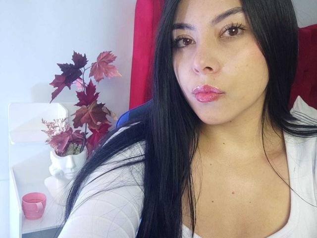 SophiaRamirez has long black hair, pink lipstick, and a white shirt, sitting in a red chair with red and brown leaves in a white vase. Boobs slightly visible.