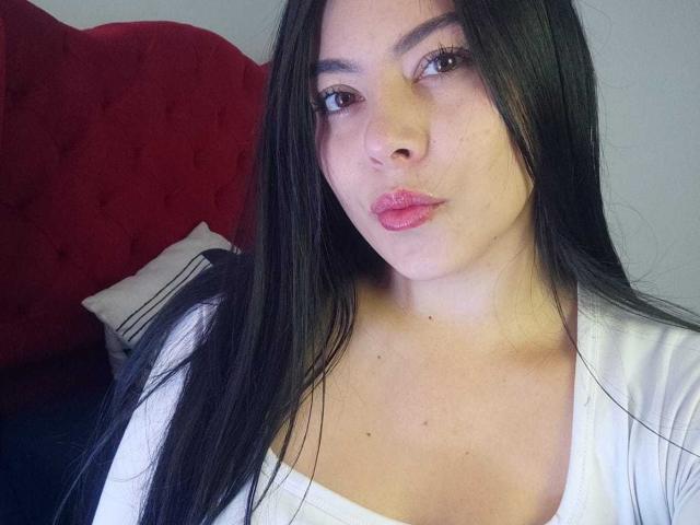 SophiaRamirez with long black hair, pink lipstick, and a white top, pouting her full lips, showing her small tits, and a cute face in a selfie.