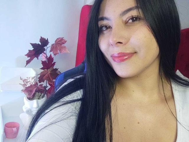 SophiaRamirez with long black hair, brown eyes, pink lips, white top, sitting in red chair, smiling, pink candle, red and purple leaves in white vase on left.