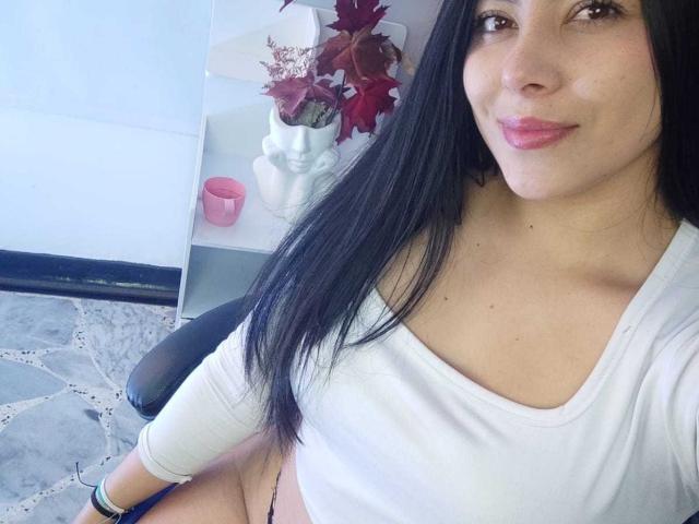 SophiaRamirez with long black hair, white top, and pink lips, showing her small tits, sitting with black chair, and pink candle in background. Her pussy is partially visible.
