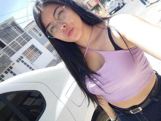 SophiaRamirez with long black hair, glasses, and pink top, showing her small tits in a purple halter, standing by a white car, pouting with her pink lips.