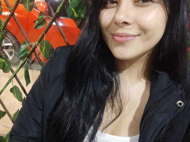 SophiaRamirez with long black hair, wearing a black jacket, smiling, showing her small tits in a white top, standing by a green plant, next to a red car.
