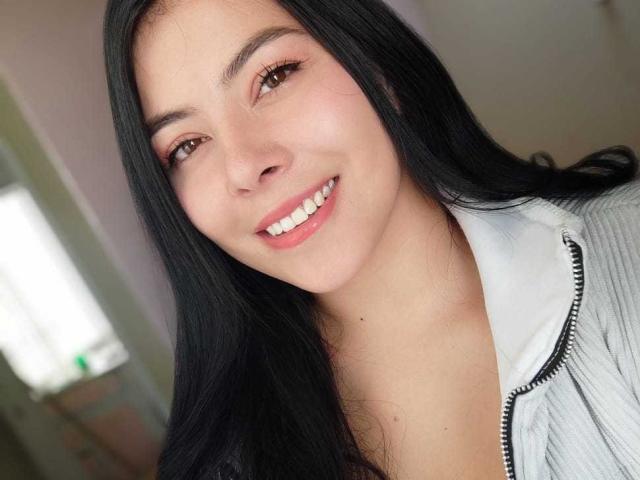 SophiaRamirez with long black hair, brown eyes, and a smile showing white teeth. She's wearing a gray zip-up shirt with a white collar. Her skin is light.