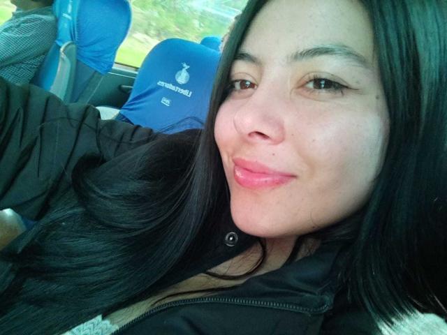 SophiaRamirez has shiny black hair, light skin, and pink lips, wearing a black jacket, sitting in a car with blue seats, smiling slightly at the camera.