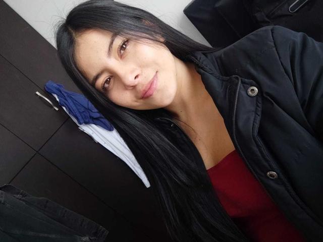 SophiaRamirez has long black hair, brown eyes, and a slight smile. She's wearing a black jacket over a red top, with a blue and white shirt hanging behind her.
