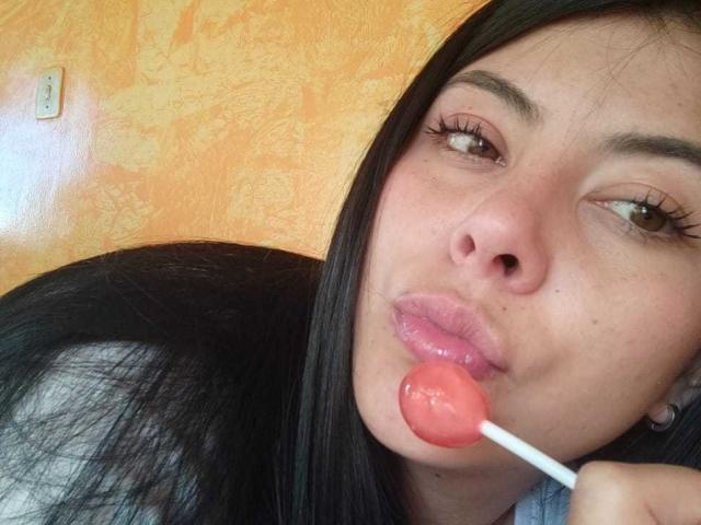 SophiaRamirez with long black hair, brown eyes, and pink lips, sucking on a pink lollipop, her face slightly tilted, showing a bit of her nose and upper lip.