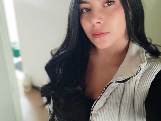 SophiaRamirez with long black hair, light skin, and pink lips, wearing a white zip-up vest over a black top, showing subtle makeup and a calm expression.