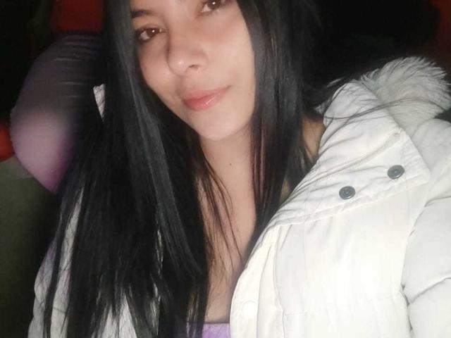 SophiaRamirez with long black hair, wearing a white puffy jacket, slightly smiling, showing pinkish lips, and brown eyes, in a dimly lit setting.